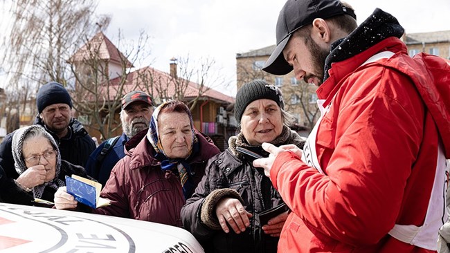 An aid worker in a red coat interacts with older adults lined up near him