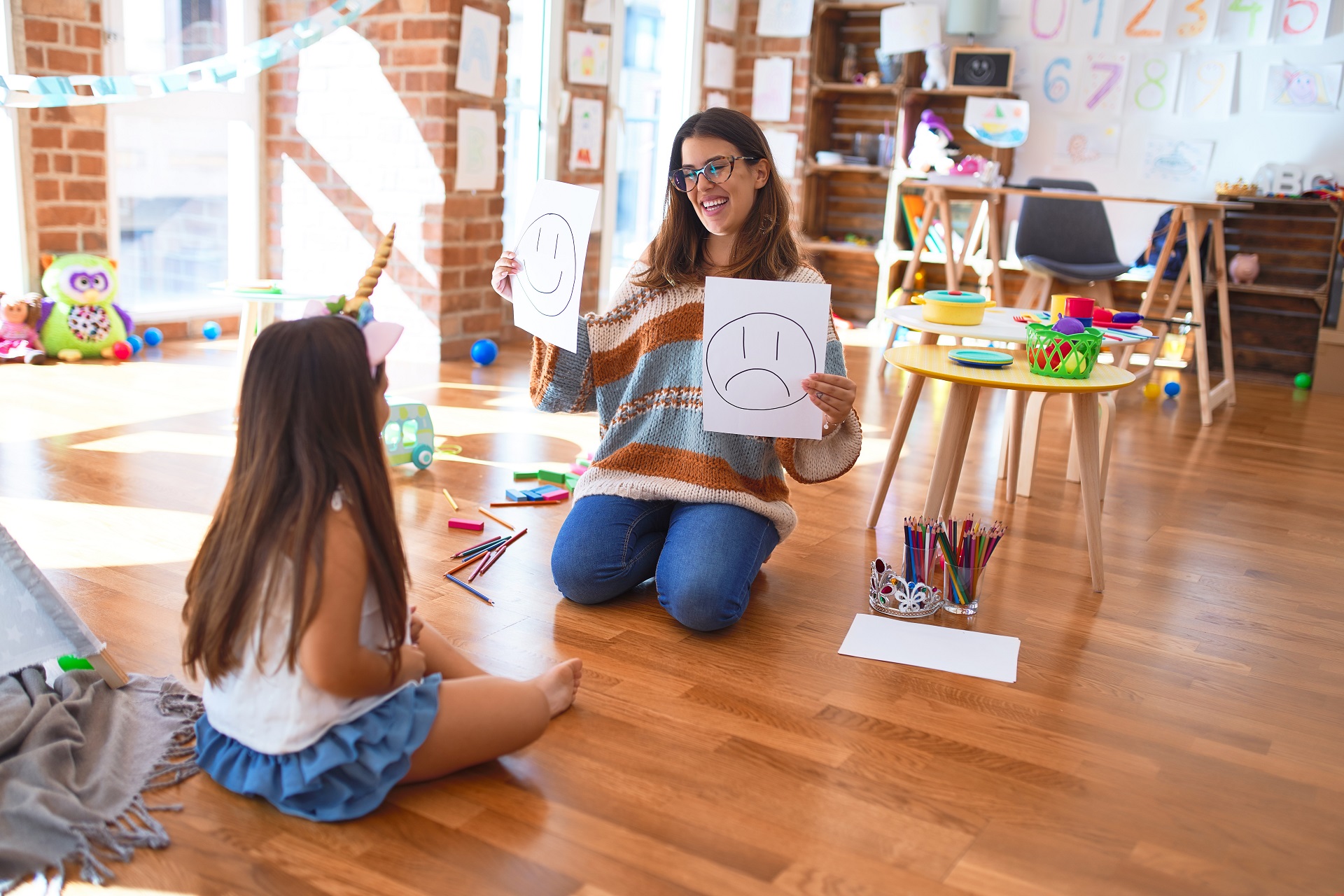 A smiling adult in a colorful, kid-friendly space holds two pieces of paper up to a child, one with a smiling face drawn on it and the other with a frowning face