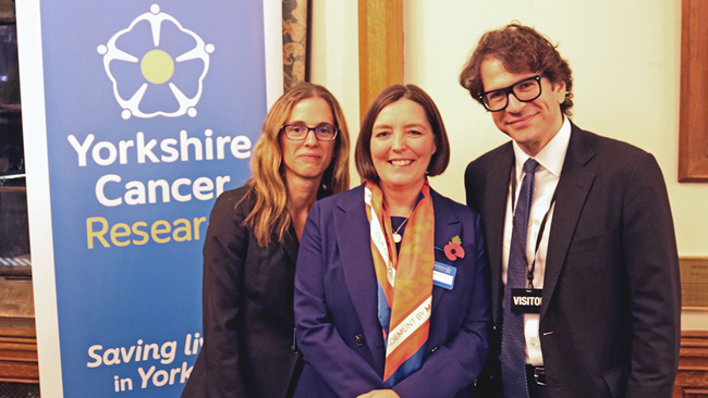Three people in business attire pose in front of a banner that reads "Yorkshire Cancer Research"
