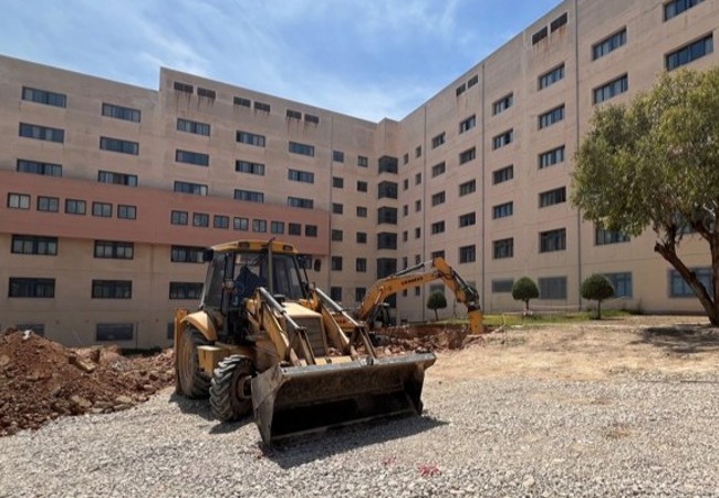 Construction equipment in a courtyard beside the Attikon Hospital building under a clear sky