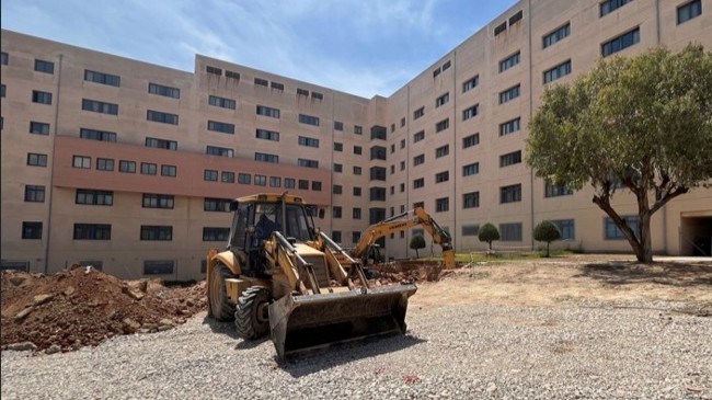Construction equipment in a courtyard beside the Attikon Hospital building under a clear sky
