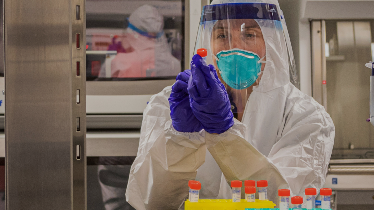 A scientist in a mask, face shield, and white coverall examines a vial