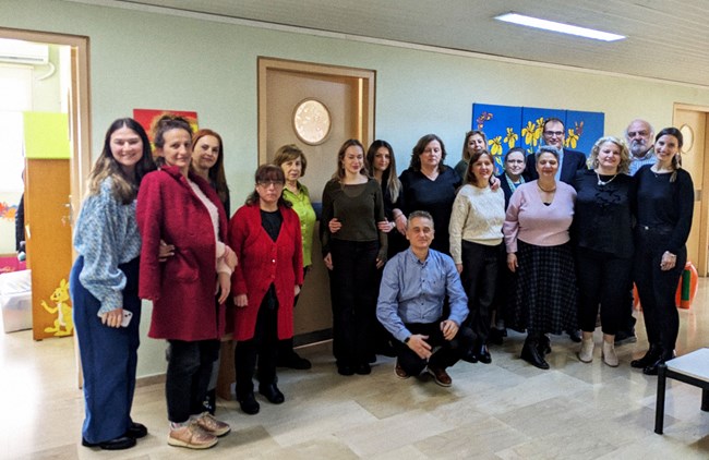 A group of 17 people stand in the corridor of a building, side by side, smiling for the camera.