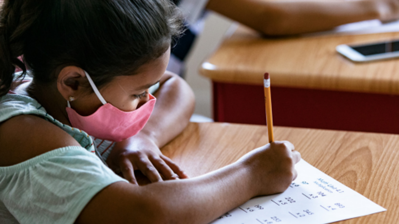 A child in a facemask writes with a pencil on a math worksheet